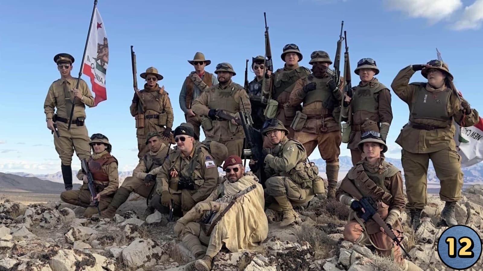 Quinze pessoas em trajes militares variados posam em terreno rochoso com montanhas ao fundo. Algumas seguram rifles; duas exibem bandeiras brancas com urso e texto ‘N. California’. Equipamentos incluem capacetes, óculos, coletes e cintos táticos.