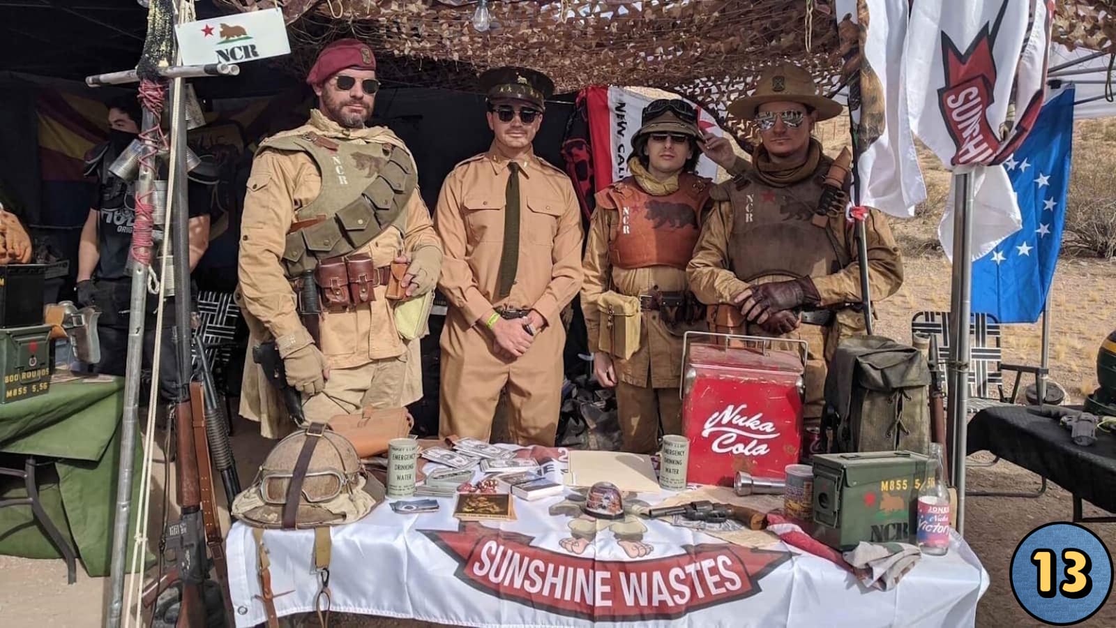 Quatro pessoas fantasiadas como facção NCR posam atrás de mesa com itens temáticos: cooler vermelho, caixas militares, capacete com óculos, latas de água de emergência, dinheiro cenográfico e banner ‘Sunshine Wastes’. Bandeiras e rede camuflada compõem o fundo em ambiente externo.