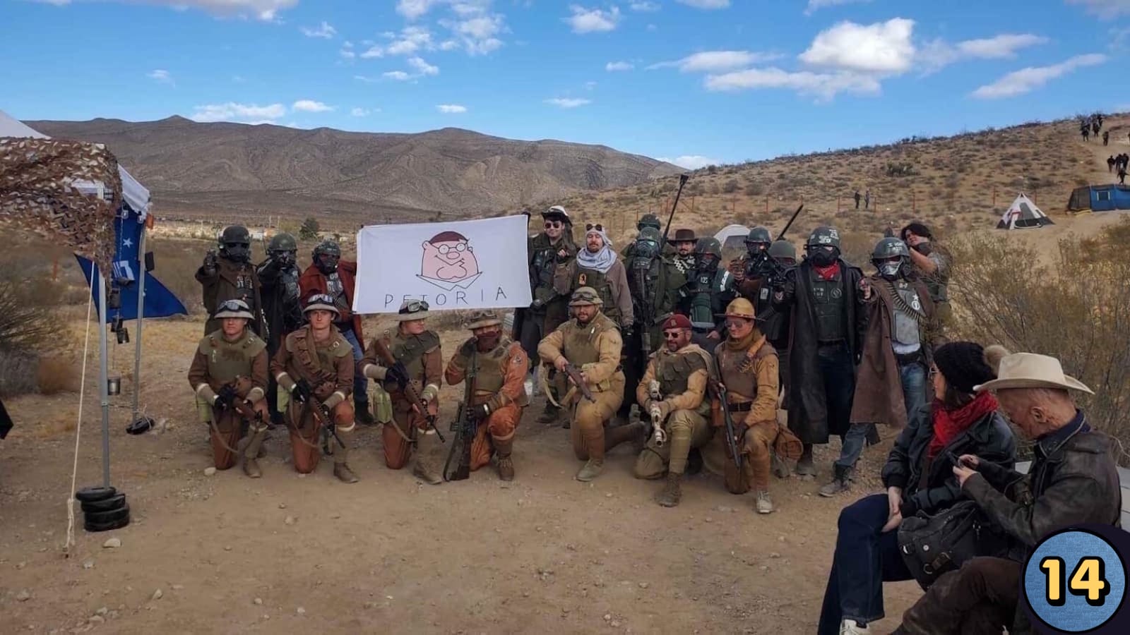 Grupo em cenário desértico com montanhas ao fundo segura bandeira branca com desenho de personagem e texto ‘Petoria’. Alguns ajoelhados, outros em pé, usando capacetes, óculos e equipamentos táticos. Tendas e participantes ao fundo. 