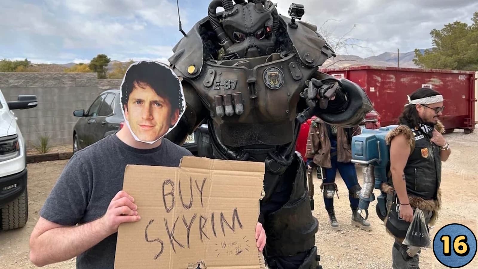 Pessoa usando máscara impressa de Todd Howard segura placa de papel ‘Buy Skyrim’. Ao lado, cosplayer em armadura T‑51b. Fundo com participantes em trajes pós‑apocalípticos, caçamba vermelha, veículos e paisagem desértica. 