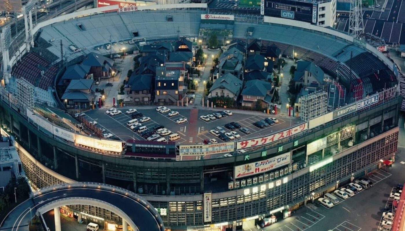 Vista aérea de estádio circular onde casas com telhados tradicionais, ruas estreitas e carros ocupam o espaço interno. Arquibancadas e anúncios em japonês permanecem ao redor, cercados por prédios urbanos e viaduto curvo.