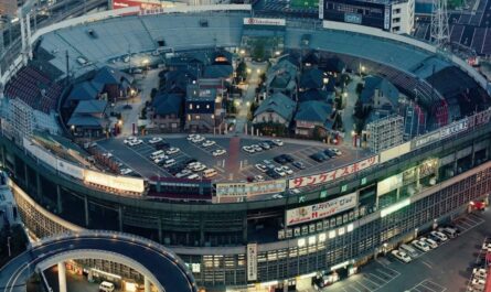 Vista aérea de estádio circular onde casas com telhados tradicionais, ruas estreitas e carros ocupam o espaço interno. Arquibancadas e anúncios em japonês permanecem ao redor, cercados por prédios urbanos e viaduto curvo.