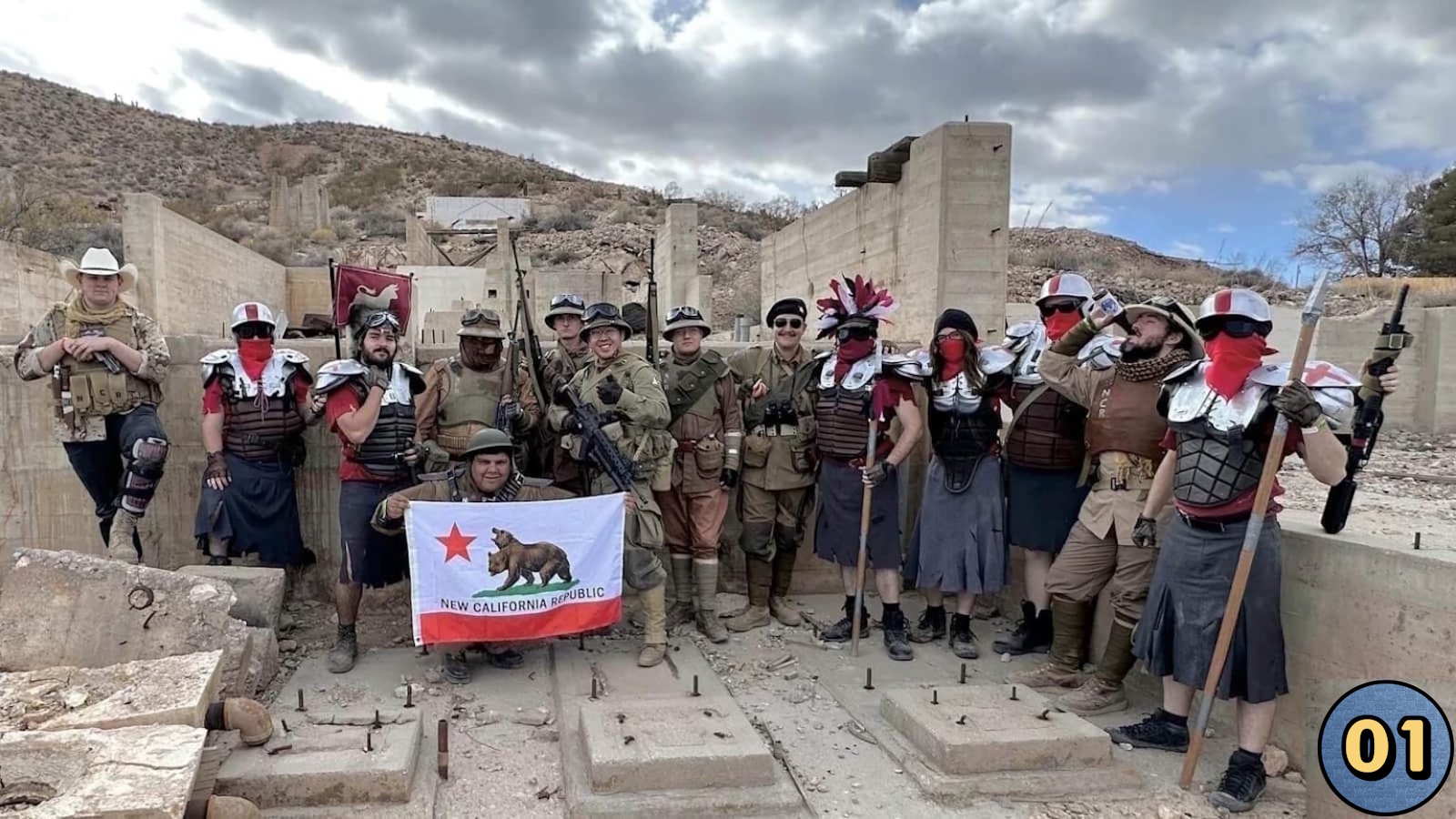 Grupo em cenário desértico com ruínas de concreto, usando armaduras e trajes inspirados em estética pós‑apocalíptica e legionária. Pessoa ao centro ergue bandeira da ‘New California Republic’; outra segura bandeira vermelha com emblema branco. Céu nublado e ambiente árido ao fundo.