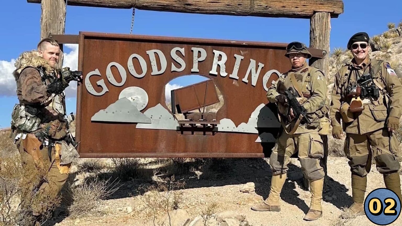 Três indivíduos com roupas militares e pós‑apocalípticas posam diante de placa metálica ‘Goodspring’ com desenho de montanhas e carrinho de mina. Equipamentos incluem óculos, coletes e armas. Paisagem desértica com vegetação rala.