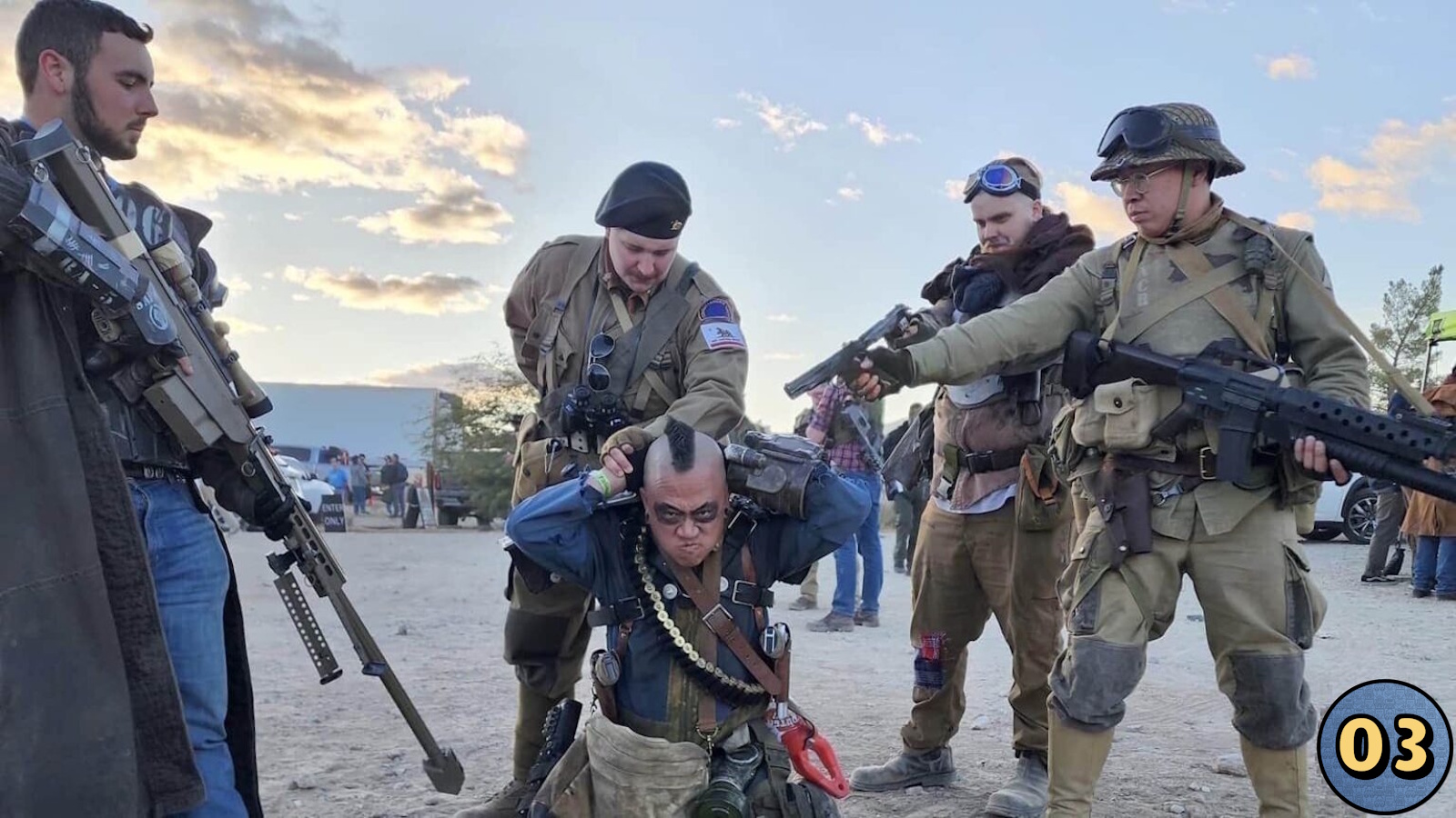 Cinco pessoas em trajes detalhados de temática pós‑apocalíptica. Figura central ajoelhada com mãos na cabeça; quatro ao redor apontam armas cenográficas. Roupas incluem capacetes, moicanos, munição e acessórios táticos. Fundo mostra área desértica com veículos e participantes. 