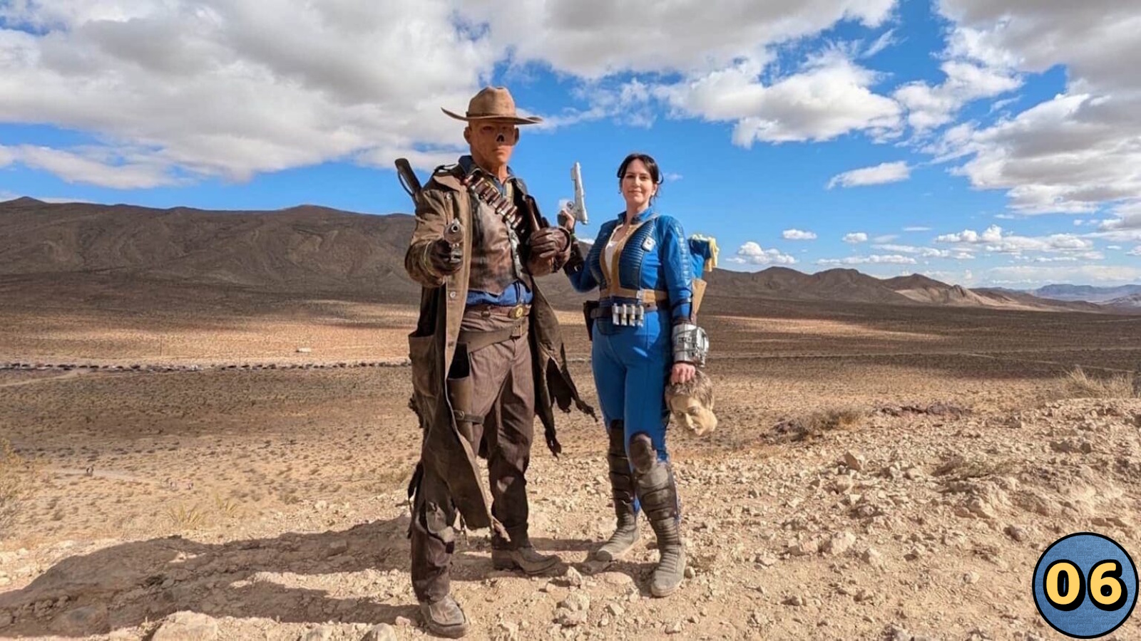 Dois cosplayers em paisagem desértica com montanhas ao fundo. À esquerda, personagem com sobretudo marrom, chapéu e arma; à direita, pessoa em macacão azul com detalhes amarelos segurando cabeça cenográfica. 