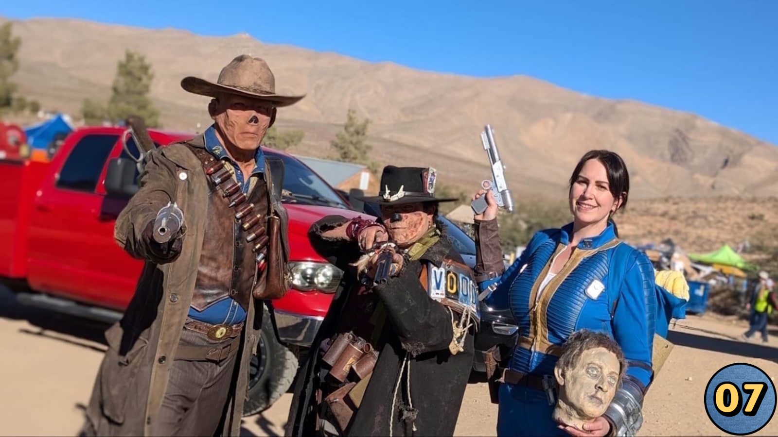 Três cosplayers em área desértica com caminhonete vermelha atrás. À esquerda, figura com chapéu, sobretudo marrom e munição no peito apontando revólver; ao centro, personagem com casaco escuro e chapéu decorado segurando arma; à direita, pessoa em macacão azul e amarelo com arma prateada e cabeça cenográfica. 