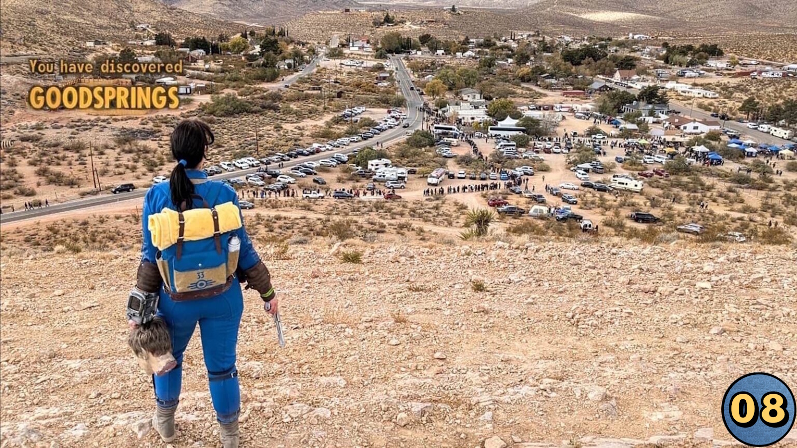 Pessoa de costas em colina rochosa usando macacão azul com detalhes amarelos, mochila e dispositivo no braço, segurando arma. Abaixo, pequena cidade com veículos, tendas e pessoas. Texto ‘You have discovered GOODSPRINGS’ aparece em amarelo. 