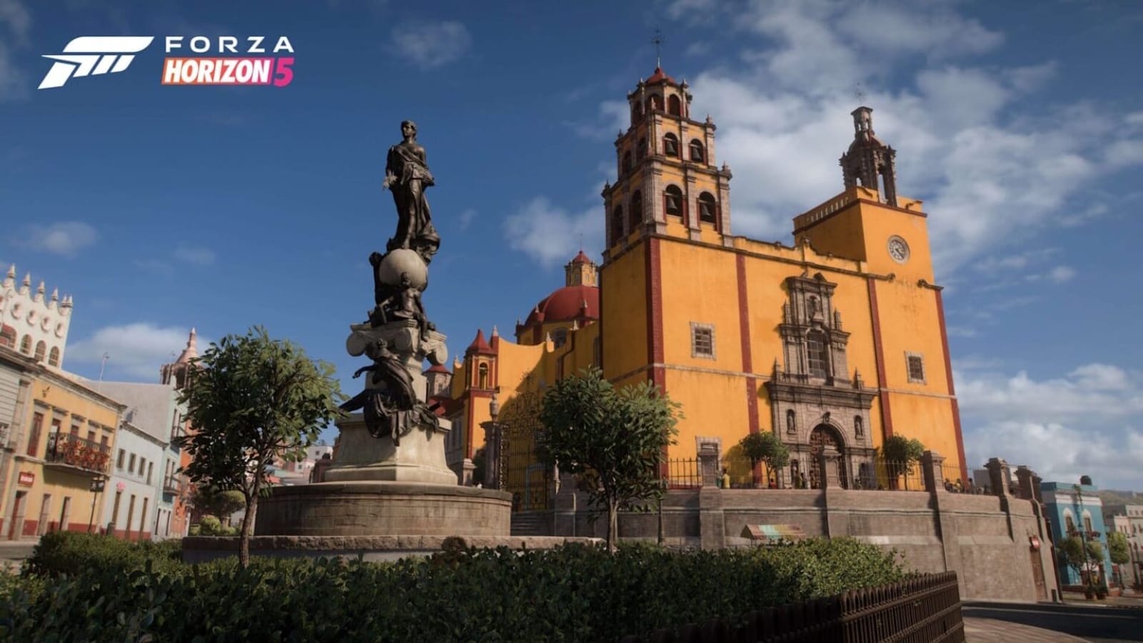 Renderização de cidade com monumento de pedra com figuras esculpidas e globo no topo. Ao fundo, igreja colonial amarela e vermelha com torres, janelas arqueadas e relógio. Vegetação ornamental ao redor e céu claro iluminando a cena. Logo ‘Forza Horizon 5’ aparece no canto superior esquerdo.