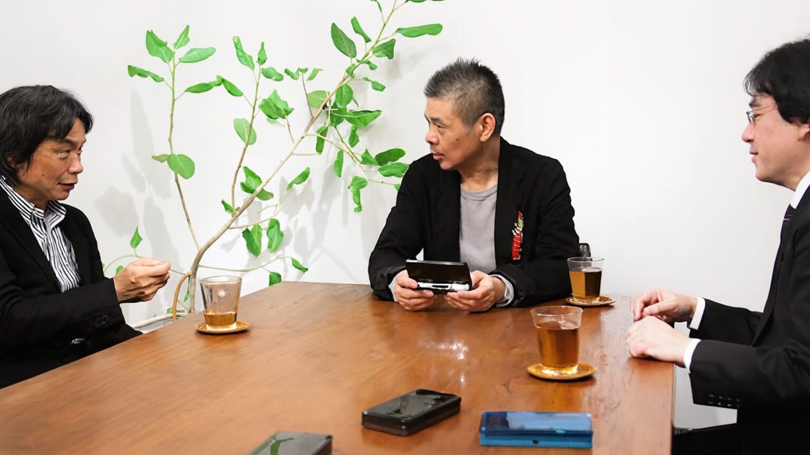 rês pessoas de terno conversando em mesa de madeira com xícaras de chá, dois celulares e console portátil, planta ao fundo.