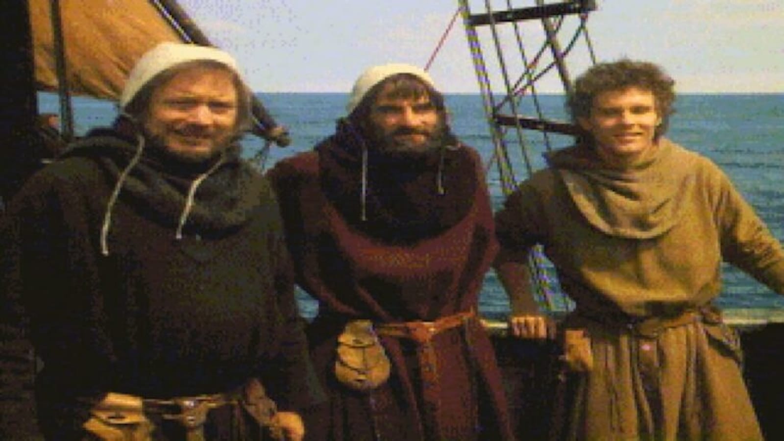 Três pessoas vestidas com roupas de estilo medieval, túnicas, cintos e capuzes, estão em pé no convés de um navio de madeira. Atrás delas aparecem cordas, mastros e parte da vela, com o mar ao fundo. A cena lembra uma viagem marítima histórica ou uma reencenação medieval.