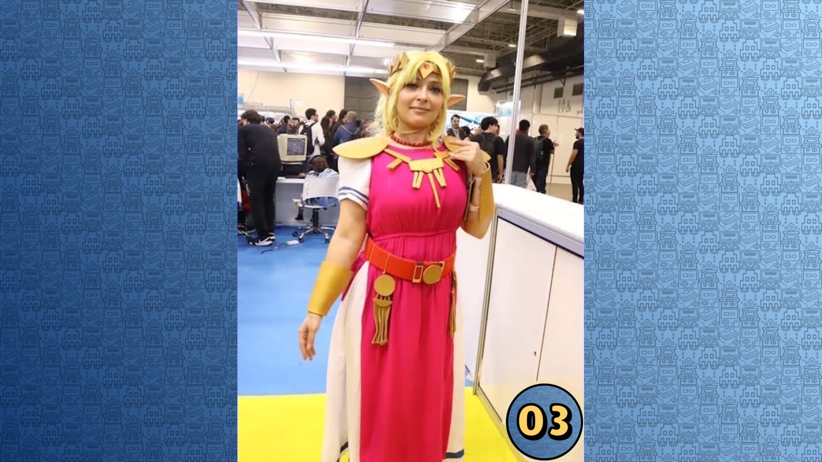 Pessoa em cosplay de Zelda com vestido rosa, armadura dourada e tiara, posando em área interna de convenção com chão azul e amarelo.