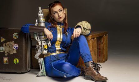 Pessoa com macacão azul e amarelo estilo Fallout sentada no chão segurando arma grande apoiada no ombro, ao lado de caixa de madeira ‘Made in U.S.A.’ e mala com adesivos.