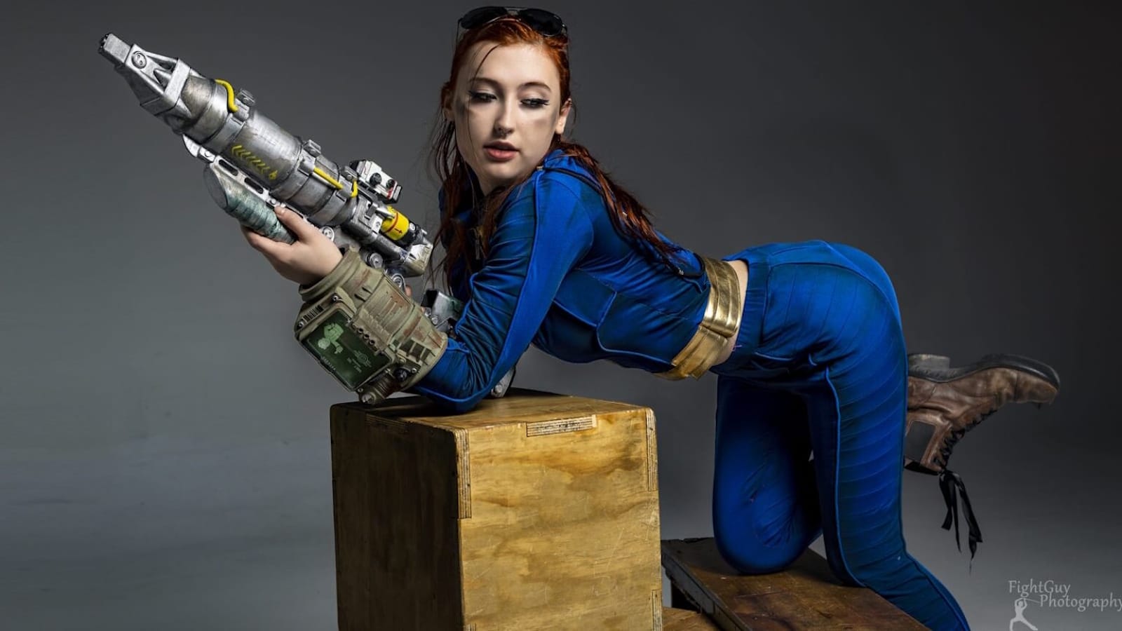 Pessoa com traje azul e dourado ajoelhada sobre caixa de madeira, segurando arma futurista. Pip‑Boy visível no braço. Fundo cinza neutro.