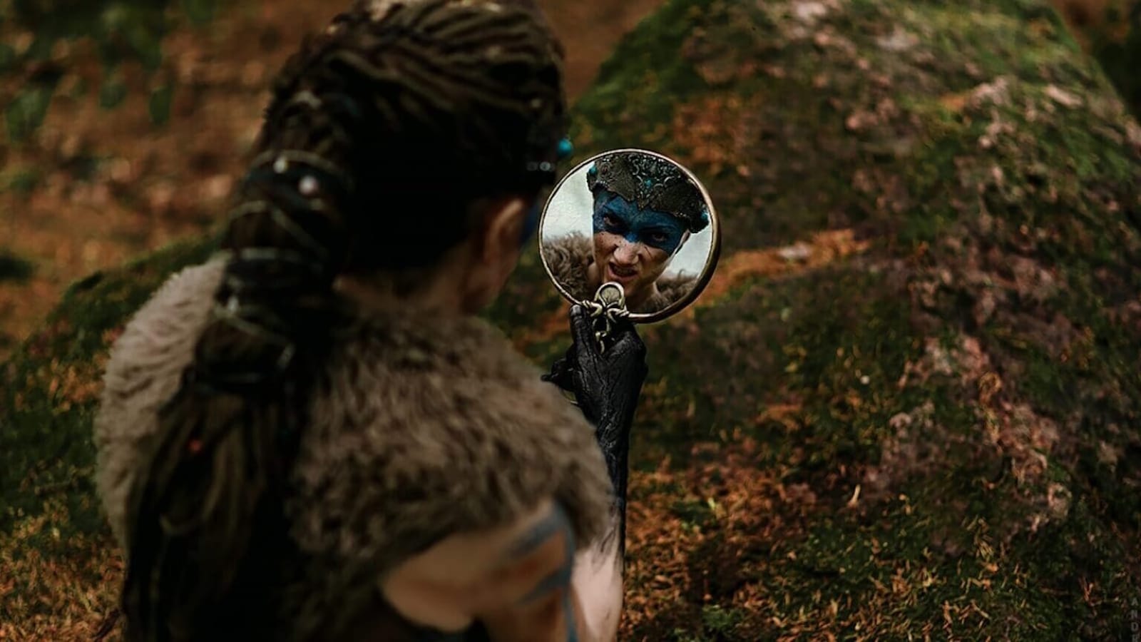 Pessoa com roupas de pele e cabelo trançado segurando espelho redondo em ambiente de floresta; reflexo mostra rosto com pintura azul e adorno decorativo.