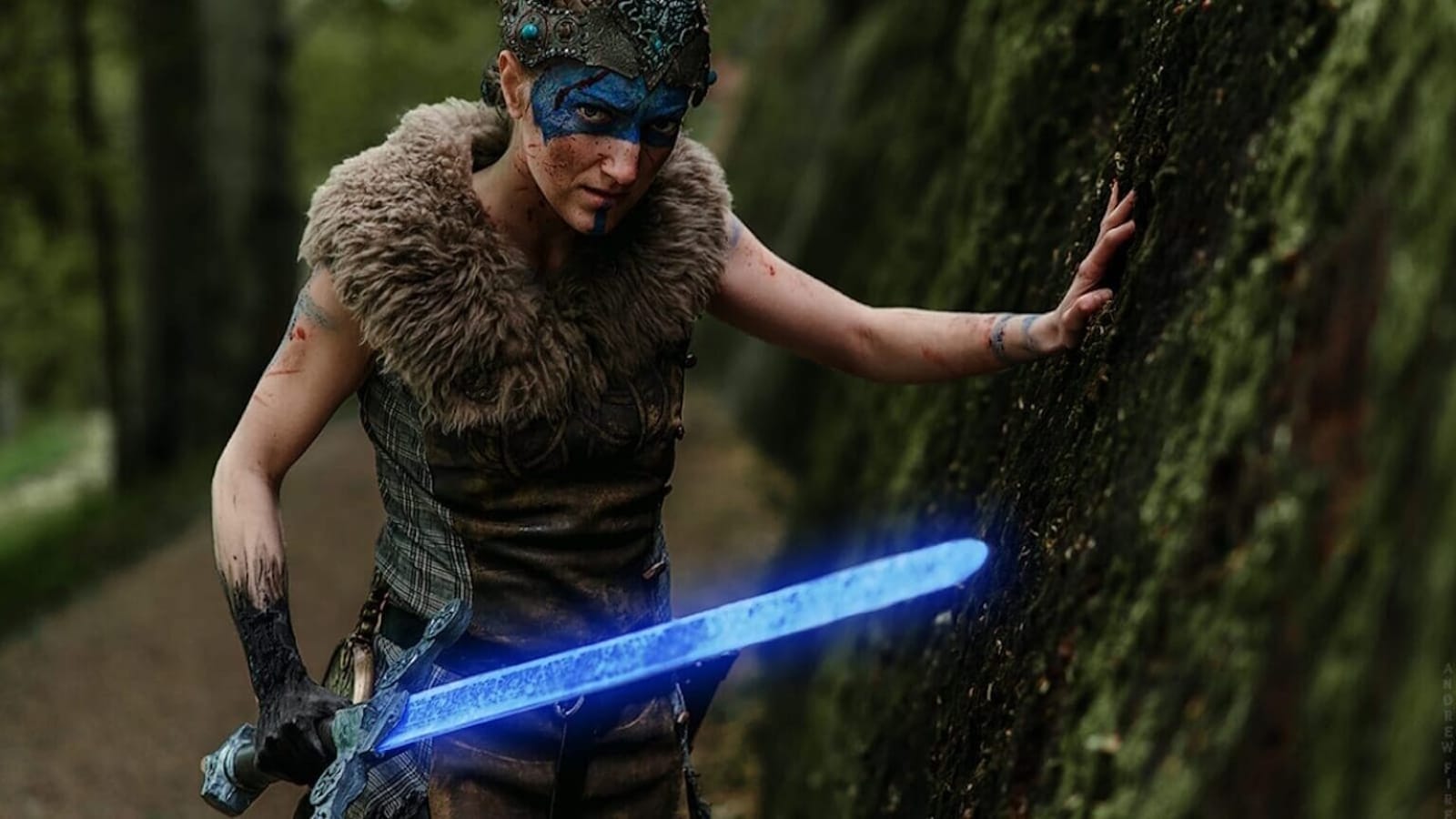 Pessoa com pintura azul no rosto e adorno decorado, usando roupa com pele, segurando espada azul luminosa e tocando parede coberta de musgo em área de floresta.