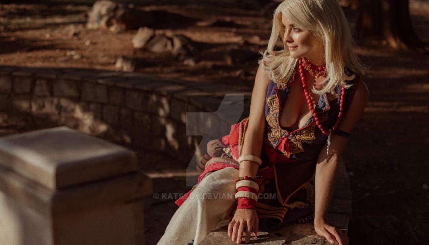 Pessoa de cabelos claros sentada sobre pedra em ambiente natural iluminado pelo sol. Usa traje elaborado com bordados, colar de contas vermelhas e várias pulseiras. Vegetação e luz dourada ao fundo.