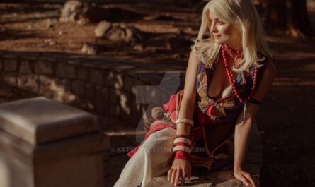 Pessoa de cabelos claros sentada sobre pedra em ambiente natural iluminado pelo sol. Usa traje elaborado com bordados, colar de contas vermelhas e várias pulseiras. Vegetação e luz dourada ao fundo.
