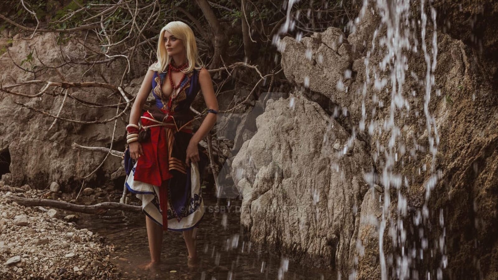 Pessoa em traje vermelho, azul e branco com padrões e contas, em pé em riacho raso próximo a paredão rochoso com pequena queda‑d’água. Galhos e folhagem compõem o cenário natural.