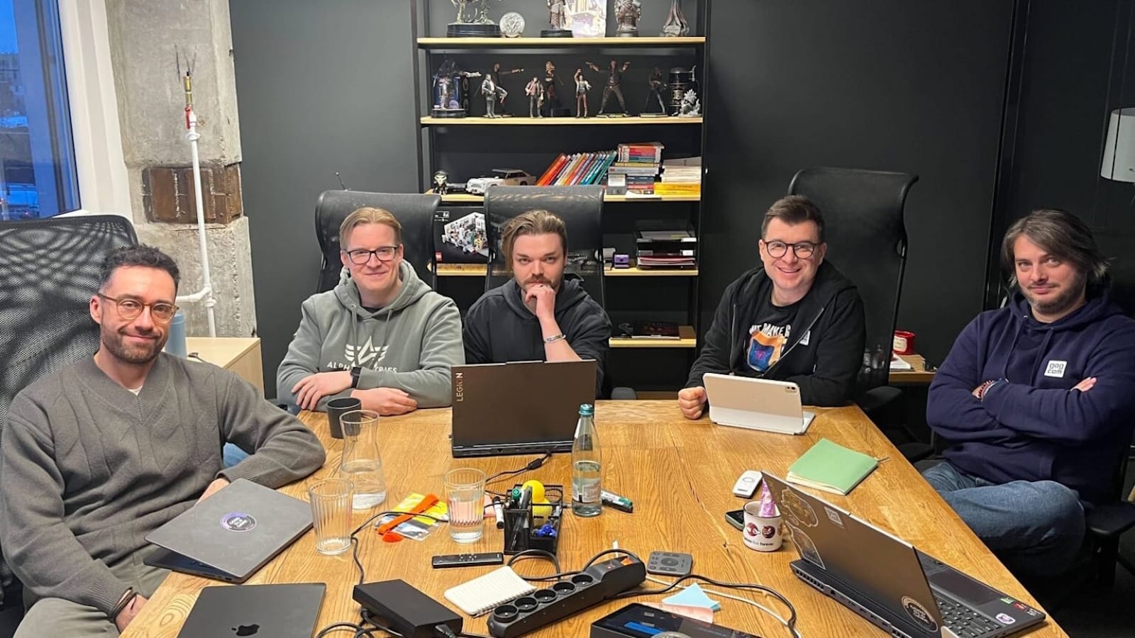 Equipe do GOG reunida ao redor de uma mesa de madeira em uma sala de reunião moderna. Cada uma tem um laptop aberto, e a mesa está cheia de itens como cadernos, canetas, garrafa de água, copos, uma régua de energia e pequenos dispositivos eletrônicos. Atrás delas há uma estante preta com livros, figuras colecionáveis e objetos decorativos.