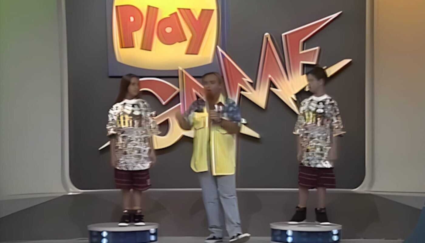 Cena de game show com três pessoas no palco: duas em plataformas circulares e uma ao centro com microfone. Painel grande ao fundo exibe logo colorido ‘Play Game’ em ambiente iluminado com paredes curvas.