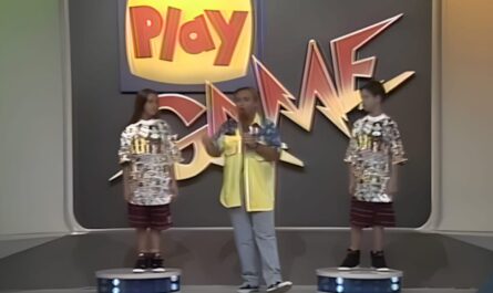Cena de game show com três pessoas no palco: duas em plataformas circulares e uma ao centro com microfone. Painel grande ao fundo exibe logo colorido ‘Play Game’ em ambiente iluminado com paredes curvas.