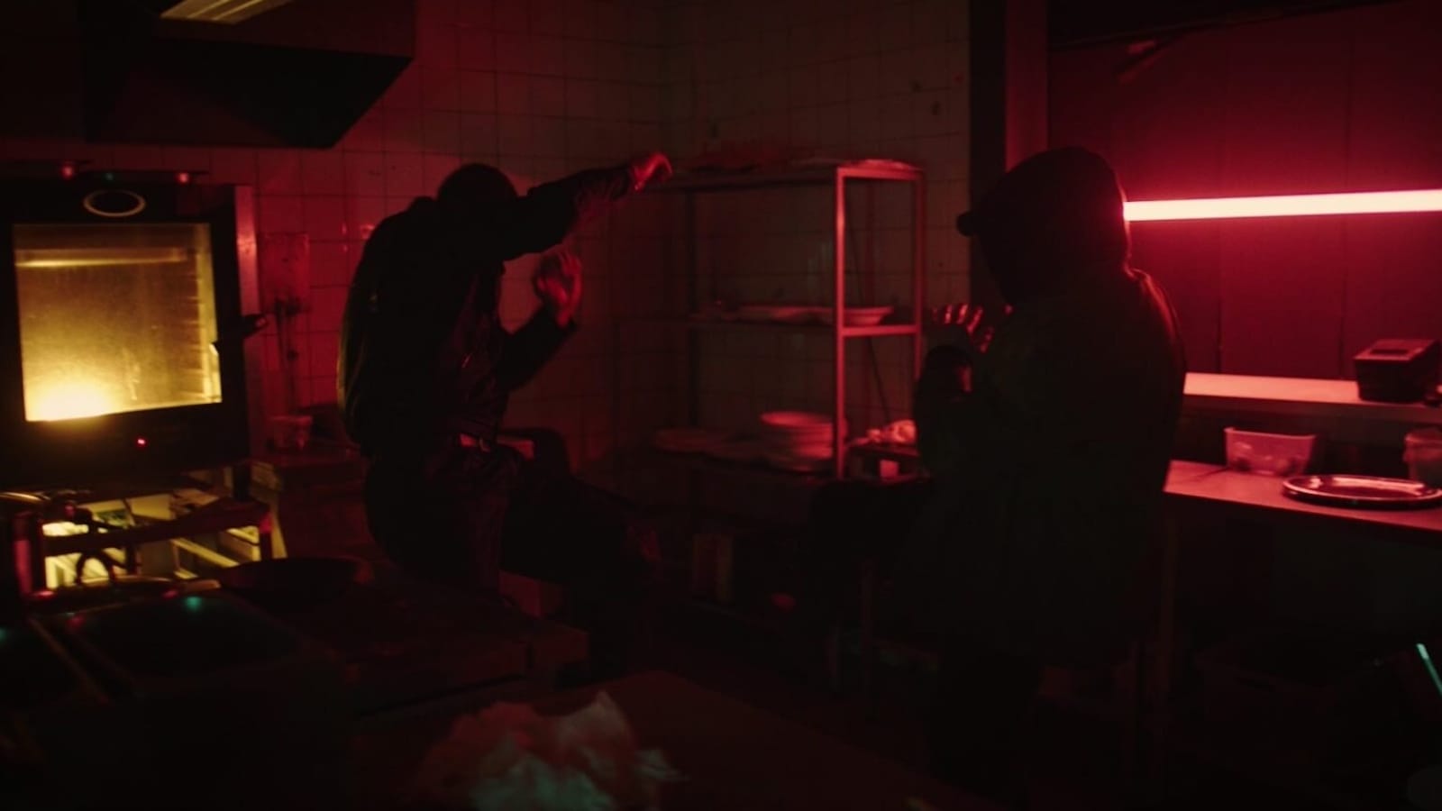 Ilustração em cozinha industrial com luz vermelha intensa. Duas pessoas se encaram: uma em pose dinâmica, outra defensiva. Ao fundo, prateleiras com pratos, bancada com utensílios e forno emitindo luz amarela.