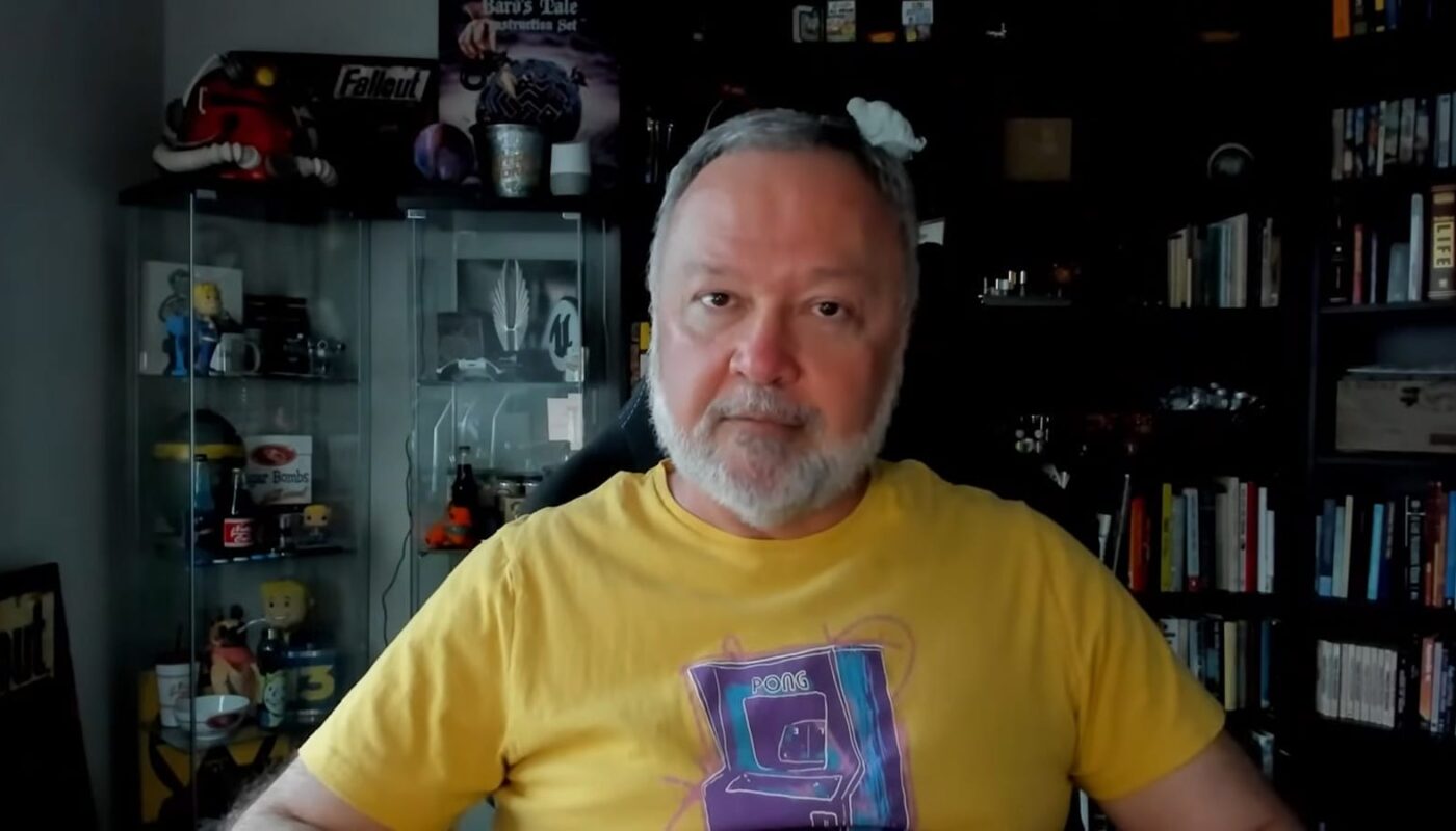Pessoa sentada usando camiseta amarela com estampa de arcade ‘PONG 2016’. Ao fundo, armário de vidro com itens colecionáveis como canecas e figuras, incluindo objetos de Fallout; pôster ‘Bard’s Tale Construction Set’ na parede e estante com livros e caixas à direita.