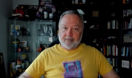 Pessoa sentada usando camiseta amarela com estampa de arcade ‘PONG 2016’. Ao fundo, armário de vidro com itens colecionáveis como canecas e figuras, incluindo objetos de Fallout; pôster ‘Bard’s Tale Construction Set’ na parede e estante com livros e caixas à direita.