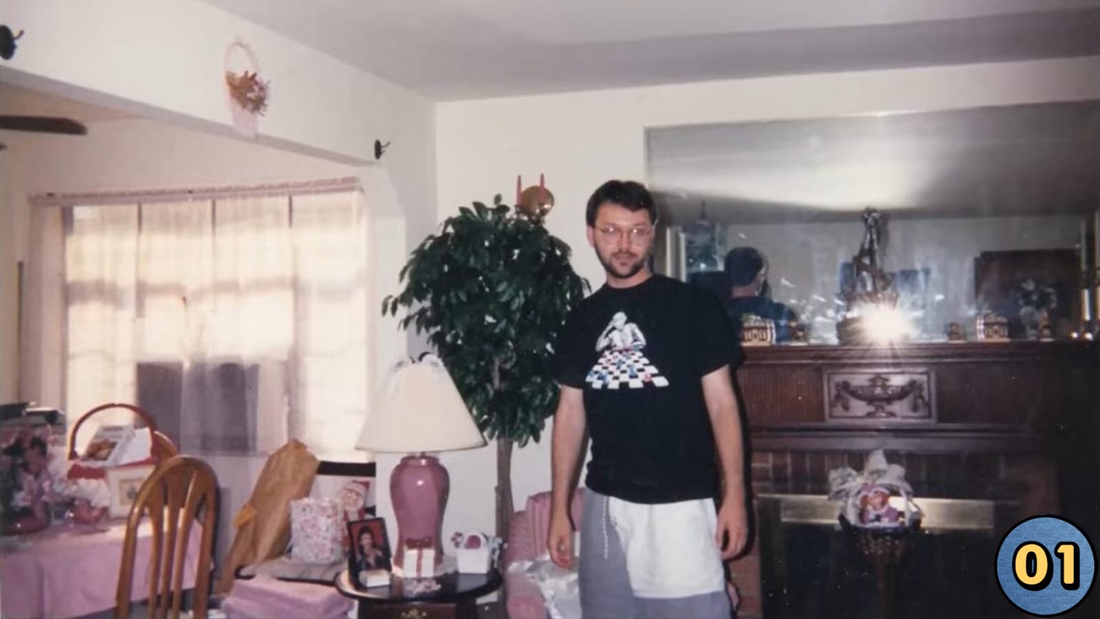 Pessoa em sala de estar com mesa de jantar coberta por toalha rosa, sacolas de presente e fotos. Há planta grande ao fundo, lareira com espelho refletindo o flash e abajur rosa sobre mesa pequena. A pessoa usa óculos, camiseta preta estampada e shorts claros. Ícone ‘01’ aparece no canto inferior direito.