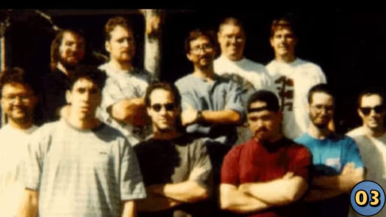 Foto de onze pessoas ao ar livre, distribuídas em duas fileiras, vestindo camisetas e alguns com óculos escuros. Fundo escuro, possivelmente sombreado. Ícone circular azul com número ‘03’ em amarelo no canto inferior direito.