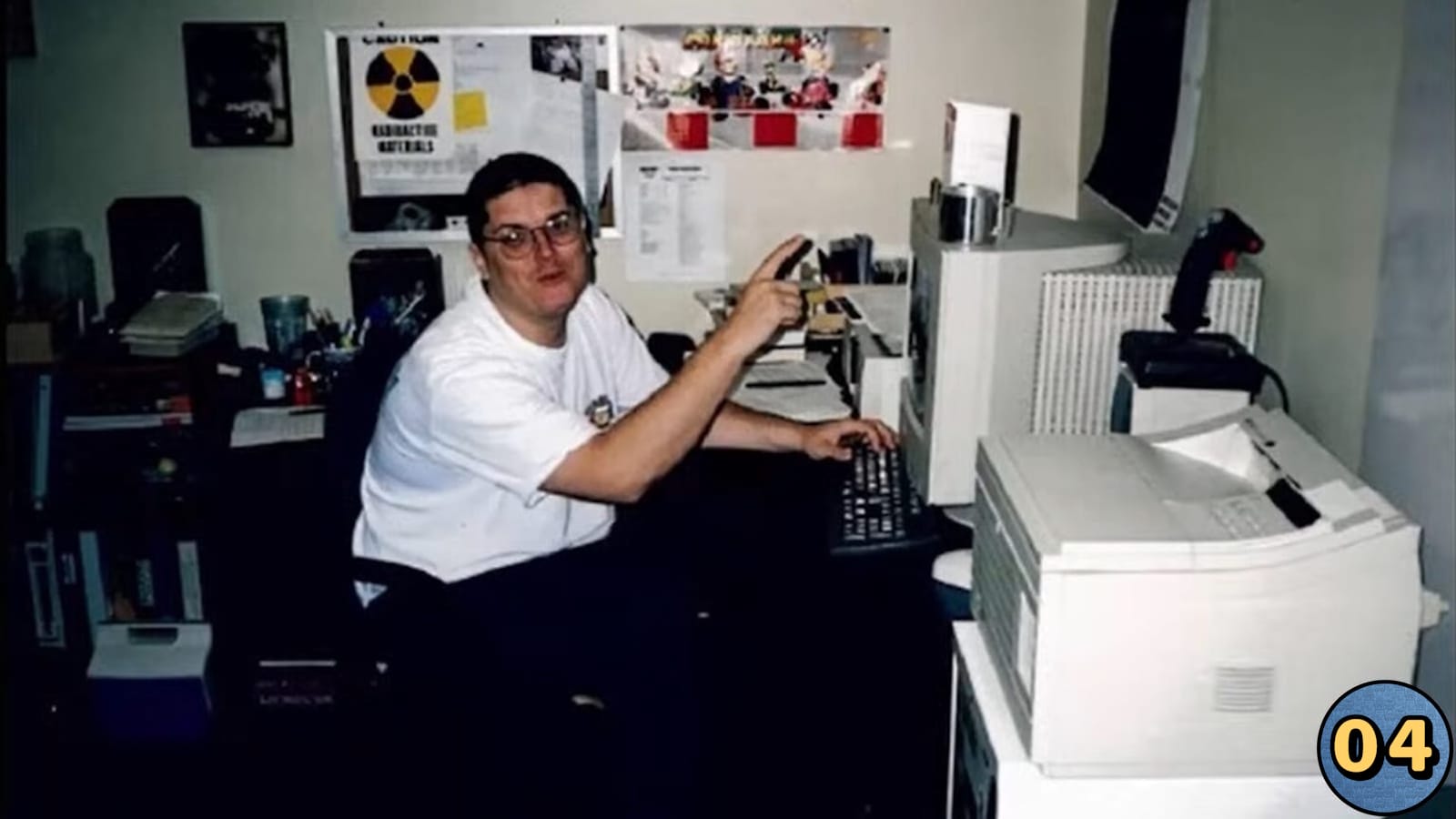 Pessoa sentada em mesa de escritório com pilhas de papéis, livros e suprimentos, apontando para monitor. Impressora grande ao lado. Parede ao fundo com fotos, papéis e placa com símbolo de radiação e texto ‘Caution Radioactive Materials’. Estilo de tecnologia remete ao fim dos anos 1990.